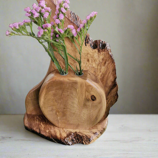 Wooden vase with a natural finish holding pink flowers on a green surface.